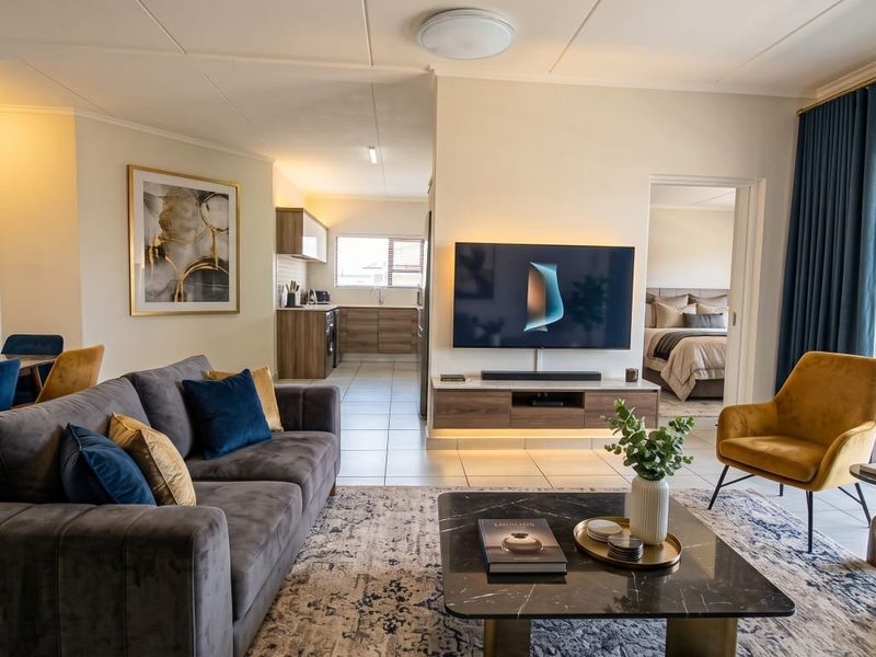 A bright open-plan living area featuring tiled floors, a modern floating media console, and seamless flow into the kitchen and adjacent bedroom.