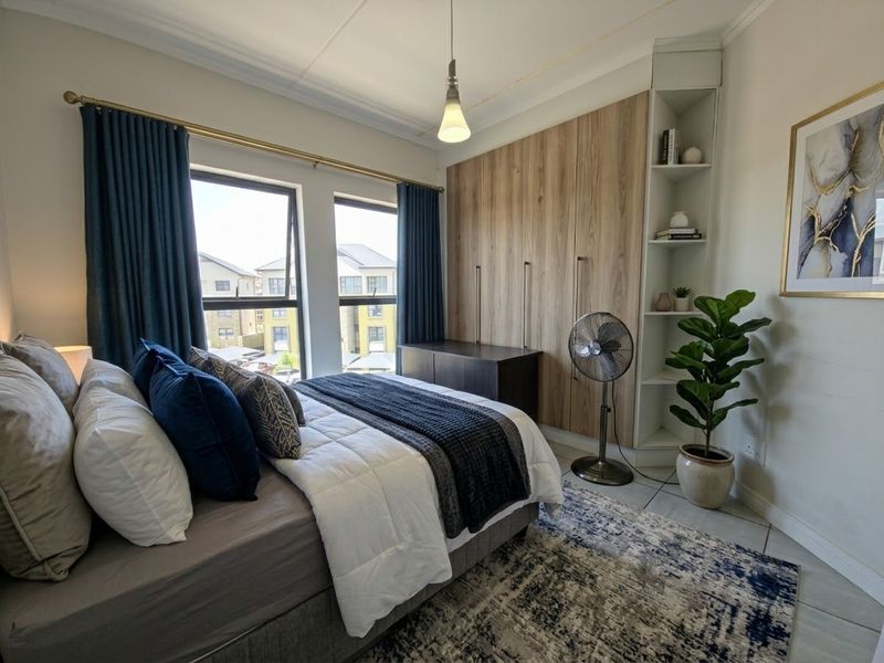 A well-maintained bedroom featuring built-in wooden cupboards, integrated shelving, tiled flooring, and large windows providing natural light.