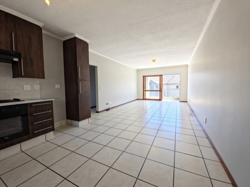 Spacious open-plan living and kitchen area featuring tiled flooring, built-in cabinetry with integrated oven and hob, and access to a balcony via sliding doors.
