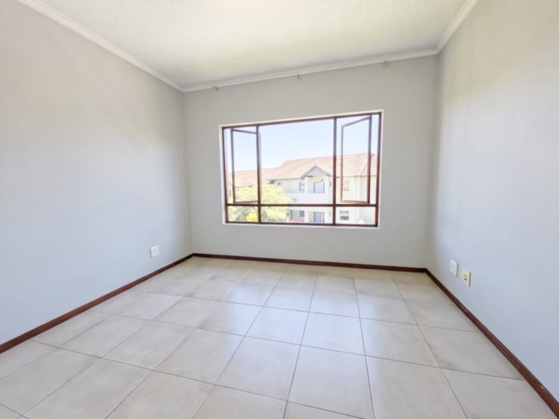A bright, empty bedroom featuring neutral tiled flooring, clean walls, and a large casement window providing natural light and a view of the complex.