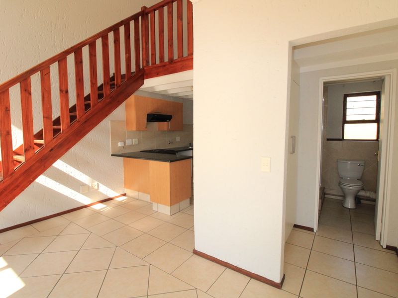 Open-plan living area featuring a wooden staircase, compact kitchen with built-in cabinetry, and an adjacent bathroom with tiled flooring throughout.