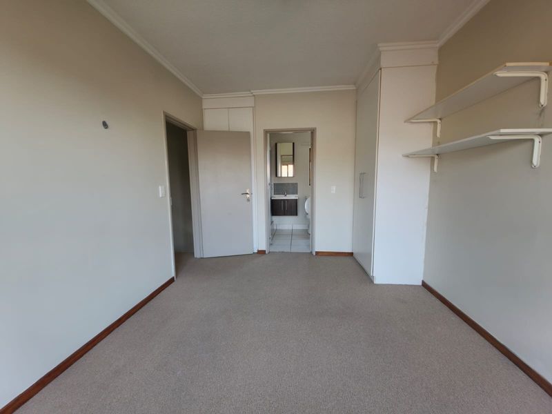 An empty bedroom featuring built-in cupboards, fitted shelving, and carpeted flooring, with an adjoining bathroom visible through the doorway.