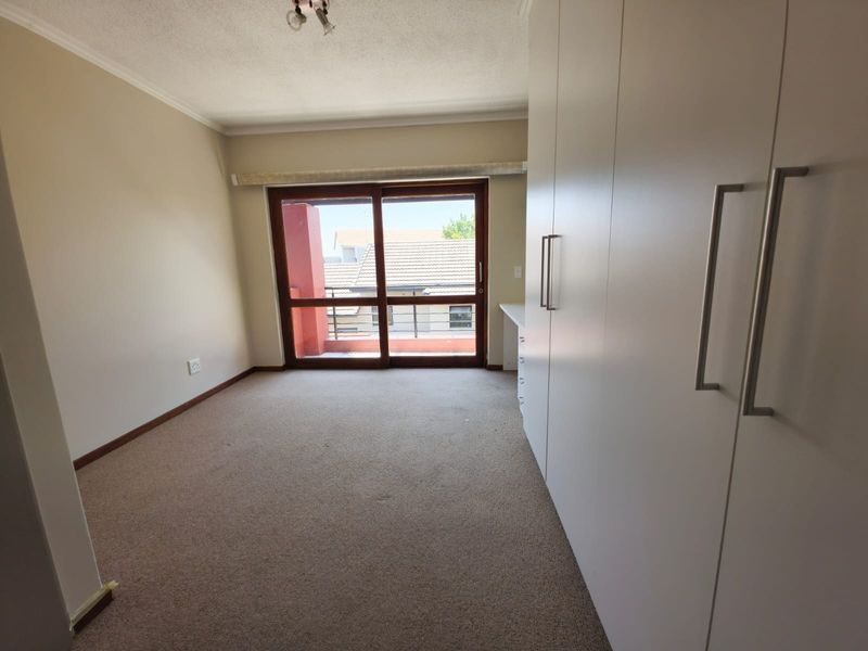 A bright, empty bedroom featuring neutral walls, carpeted flooring, and extensive built-in cupboards, with direct access to a balcony through a sliding glass door.