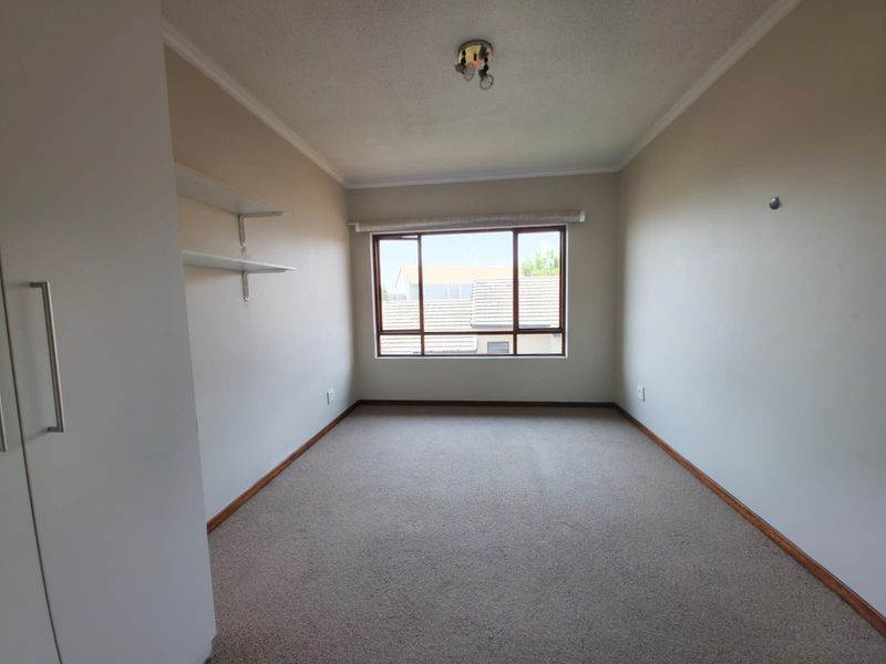 A clean, empty bedroom featuring neutral walls, carpeted flooring, built-in cupboard storage, and wall-mounted shelving with natural light from the window.