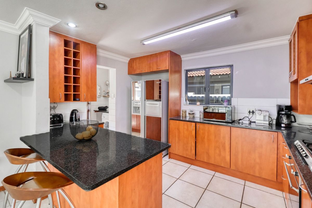 A functional kitchen featuring wooden cabinetry, granite countertops, a breakfast island, and tiled flooring, with an adjacent scullery area visible.
