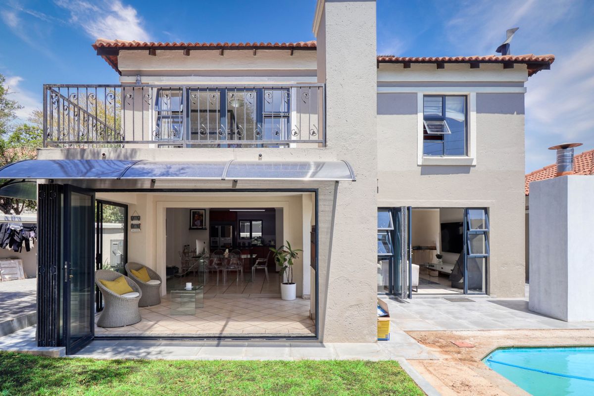 Rear view of a two-story house featuring a swimming pool, tiled patio, balcony with wrought iron railing, and large folding glass doors.