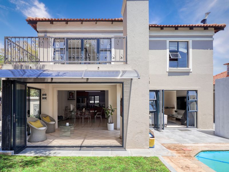 Rear view of a two-story house featuring a swimming pool, tiled patio, balcony with wrought iron railing, and large folding glass doors.