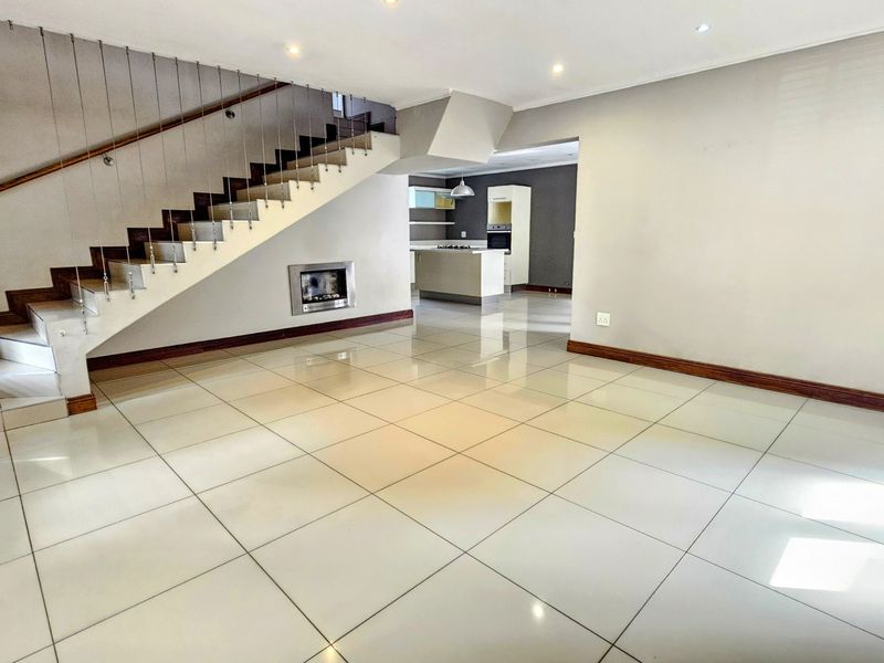 Spacious open-plan living area featuring large format floor tiles, a modern staircase with cable railing, and a built-in fireplace, flowing into the kitchen.