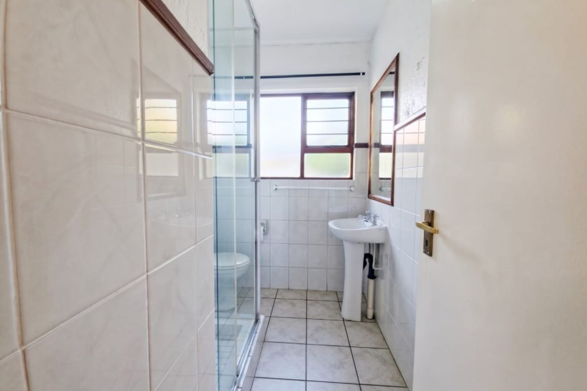 A functional bathroom featuring a pedestal washbasin, a glass-enclosed shower, and neutral wall and floor tiling.