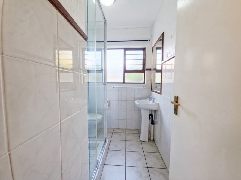 A functional bathroom featuring a pedestal washbasin, a glass-enclosed shower, and neutral wall and floor tiling.