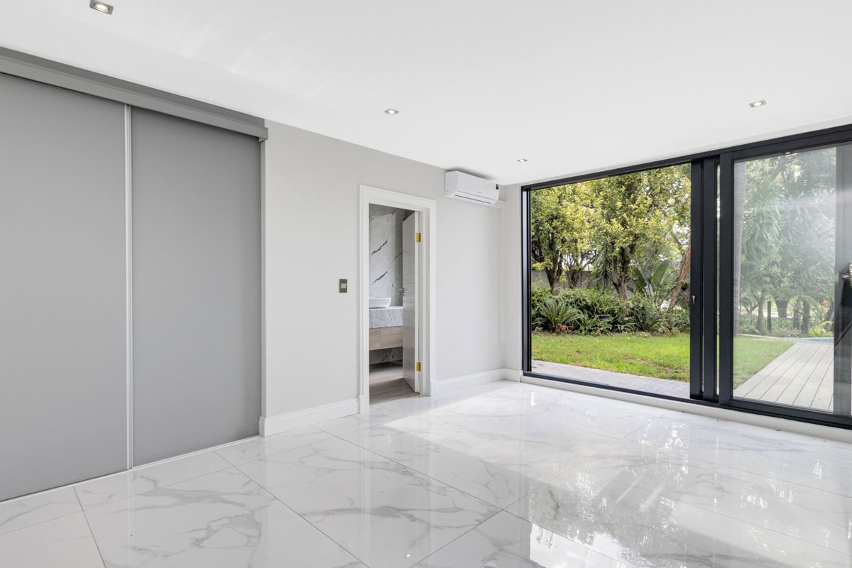 Modern bedroom featuring polished marble-look floor tiles, built-in sliding door wardrobes, split-system air conditioning, and large glass sliding doors opening to a garden view.