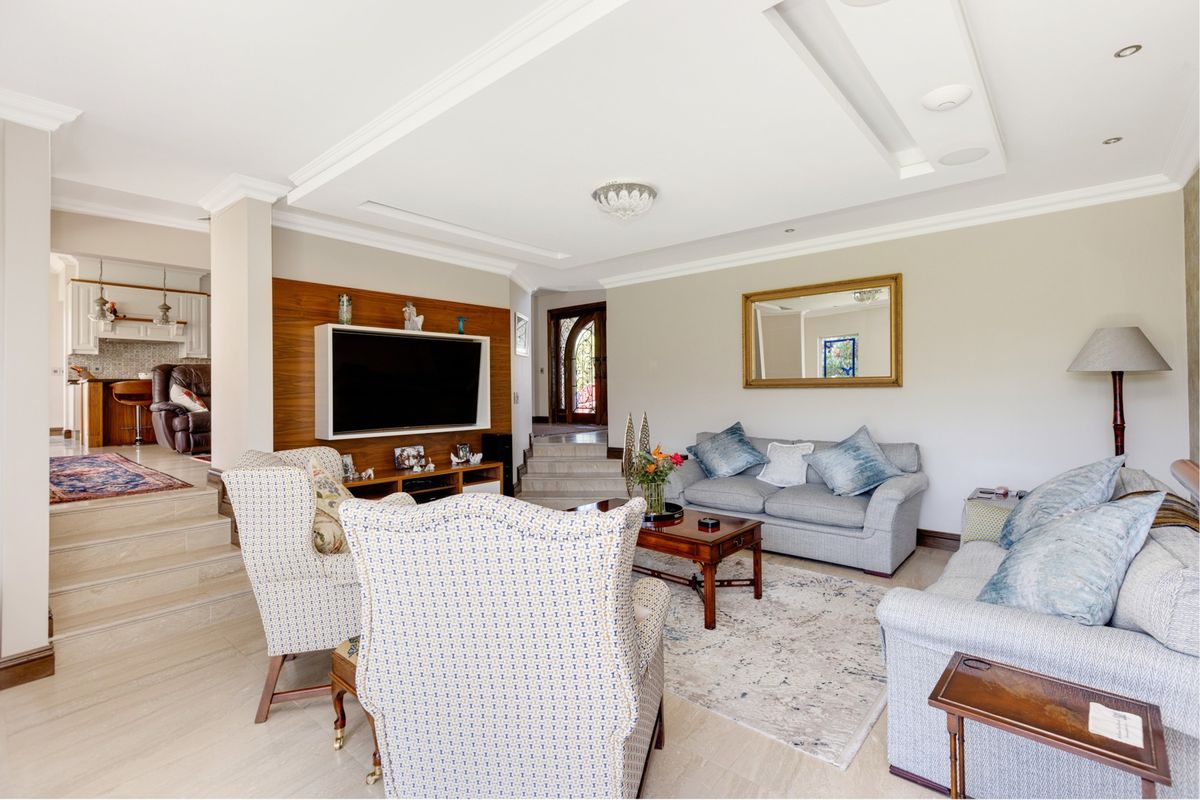 Spacious living room featuring a recessed ceiling with cove lighting, stone flooring, and a built-in media wall, with a stepped transition to the kitchen area.