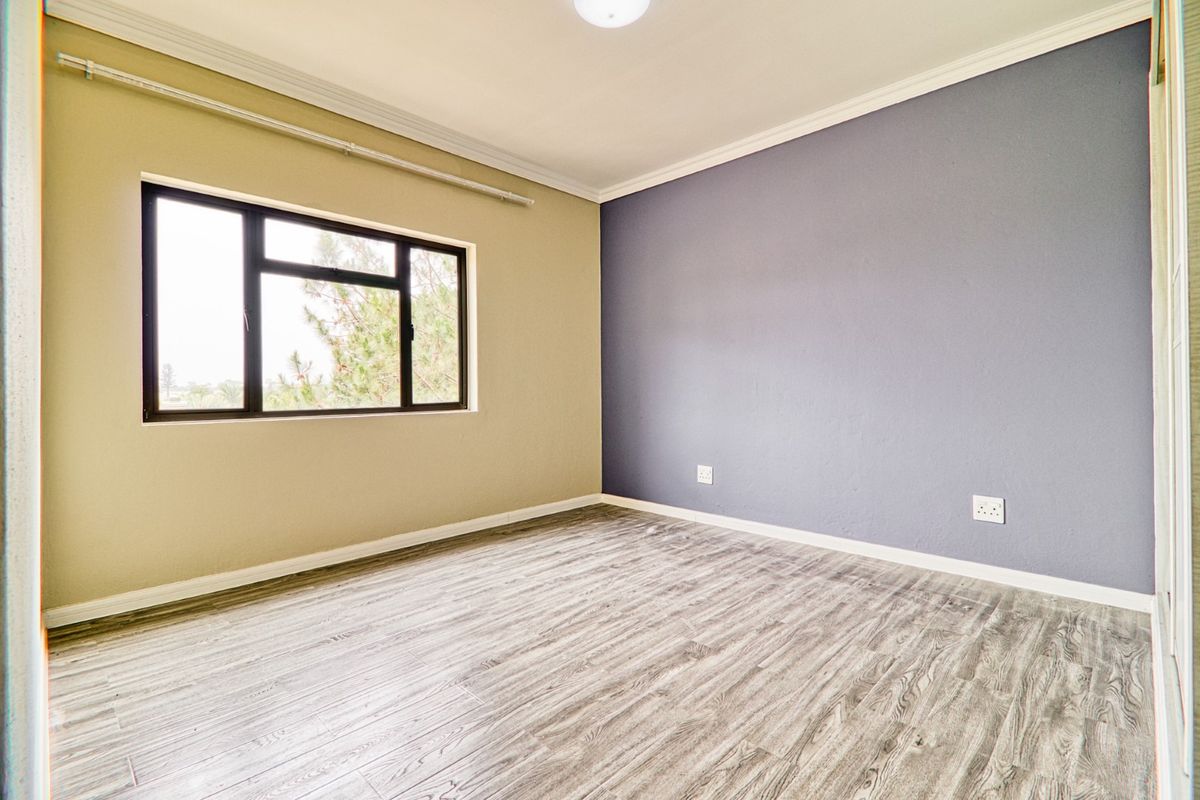 A bright, empty bedroom featuring modern wood-look laminate flooring, neutral wall tones, and a large window providing natural light.