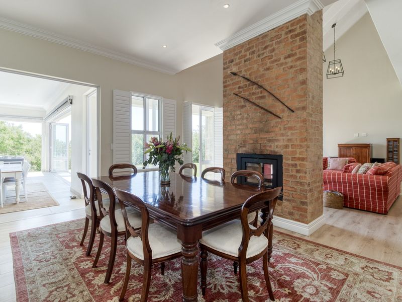 Spacious open-plan living and dining area featuring a central exposed brick fireplace, vaulted ceilings, and large windows with plantation shutters.