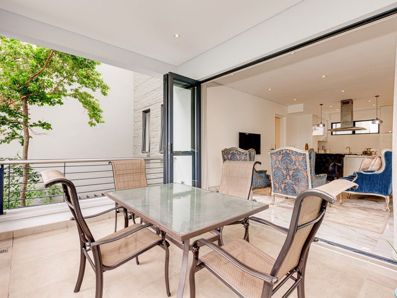 A spacious covered balcony featuring tiled flooring and metal railings, seamlessly connecting to an open-plan interior living and kitchen area through folding glass doors.