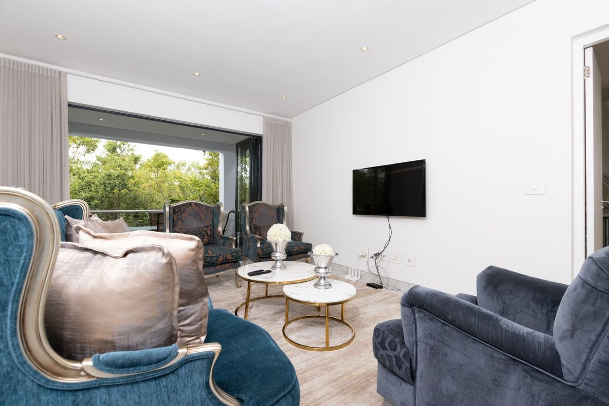 A bright living room featuring recessed ceiling lighting, wall-mounted television, and large sliding doors opening onto a balcony with lush greenery views.
