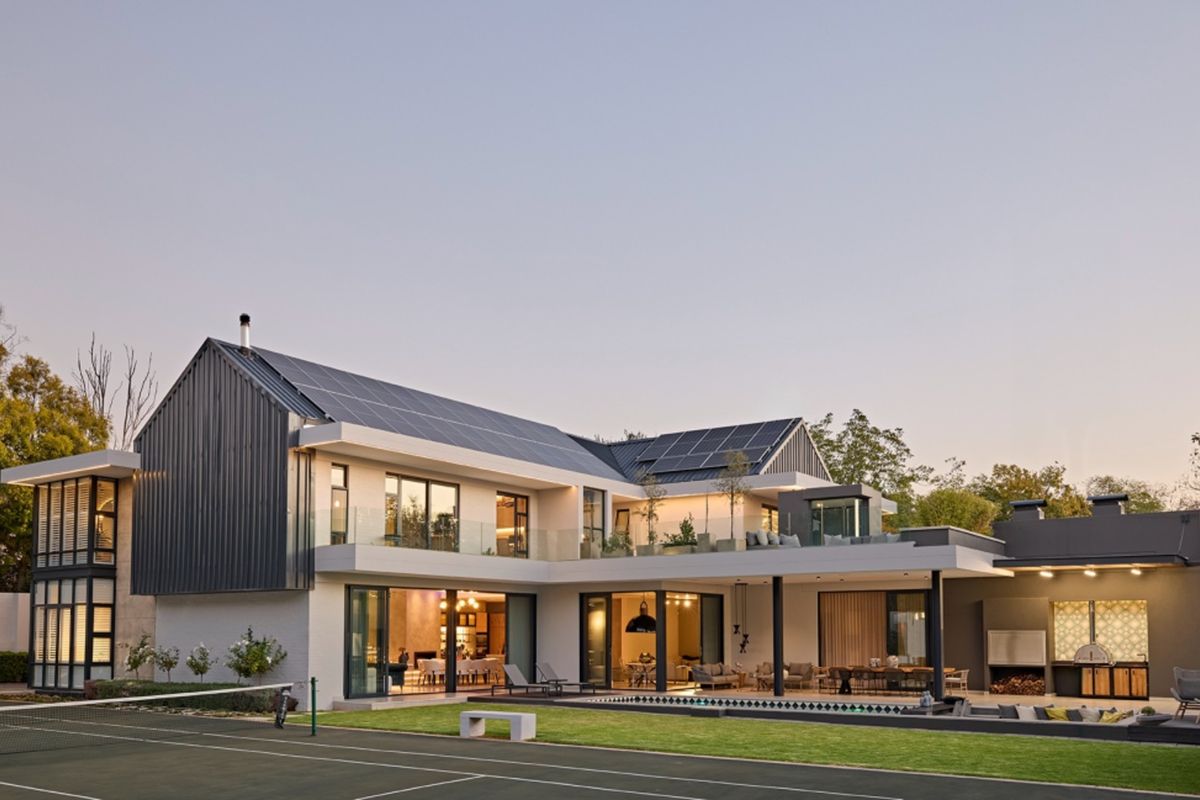 A modern luxury residence featuring a spacious rear patio, swimming pool, tennis court, and integrated solar panels on a contemporary metal roof.