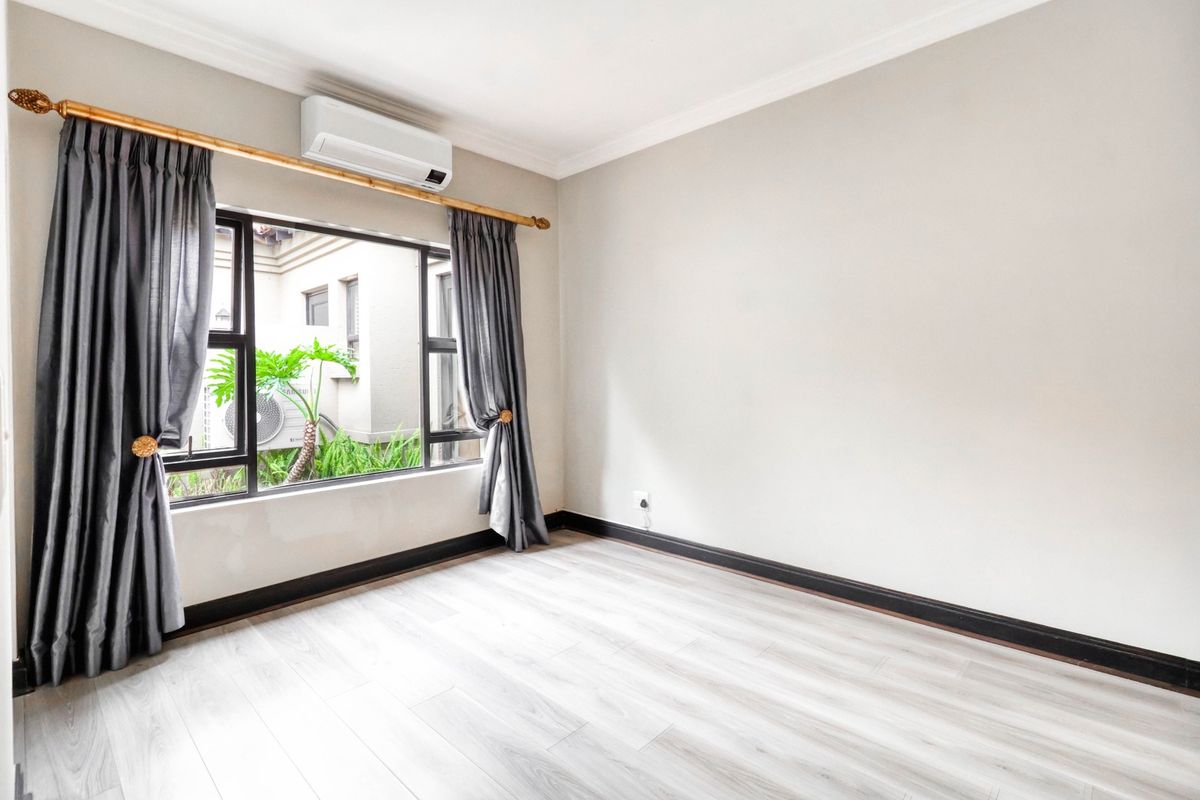A bright, empty bedroom featuring light-toned laminate flooring, neutral walls, a large window with dark curtains, and a wall-mounted air conditioning unit.
