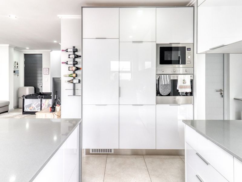 Modern kitchen featuring high-gloss white cabinetry, integrated oven and microwave, stone countertops, and a clean subway tile backsplash, flowing into an open-plan living area.