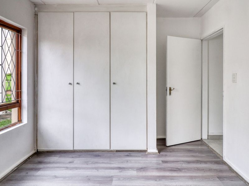 A bright, empty bedroom featuring modern grey laminate flooring and ample built-in cupboard storage, with a view overlooking the complex garden.