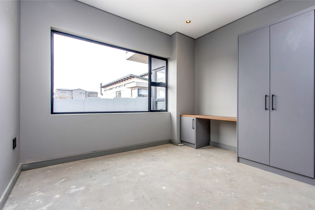 A modern, empty bedroom featuring grey built-in cabinetry, a fitted desk, and large windows, awaiting final flooring installation.