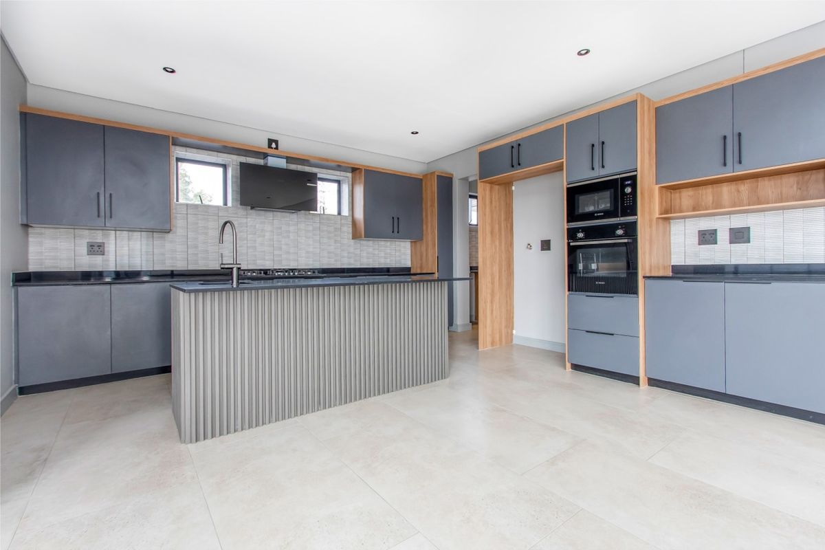 Modern kitchen featuring grey and wood-tone cabinetry, a central island with a sink, built-in appliances, and neutral floor tiles.