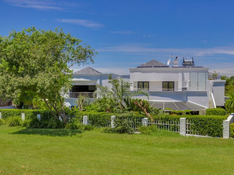 Rear view of a modern two-story house featuring a landscaped garden, metal fencing, a balcony with glass balustrades, and roof-mounted solar energy systems.