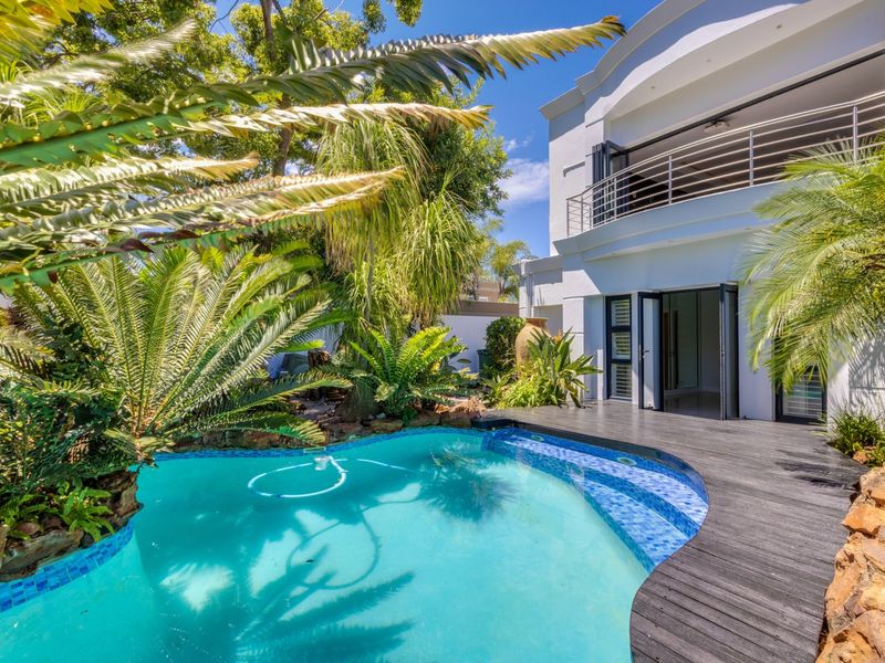 A well-maintained swimming pool area featuring timber decking, lush landscaping, and direct access to the home via bi-fold doors and an upper balcony.