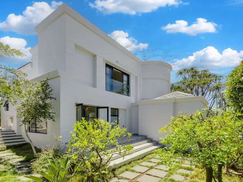 A modern white architectural exterior featuring clean lines, large windows, a glass balcony, and stone-paved steps leading into a well-maintained garden area.