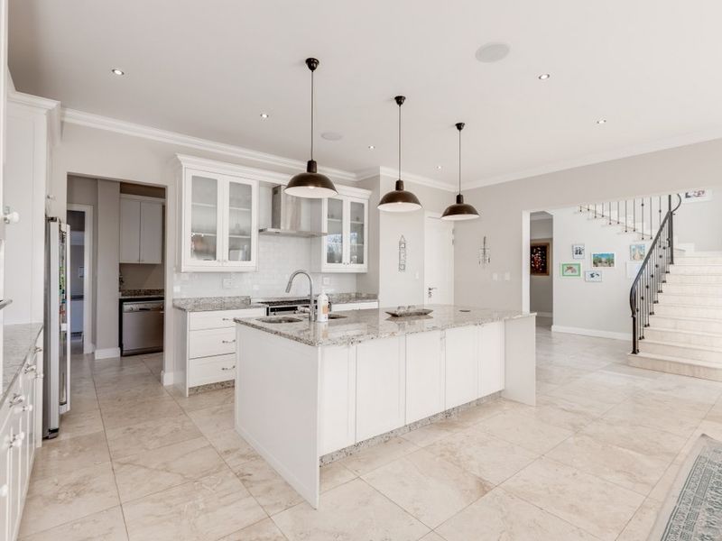 Modern kitchen featuring a central island with granite countertops, white cabinetry, stainless steel extractor fan, and tiled flooring, adjacent to an elegant staircase.
