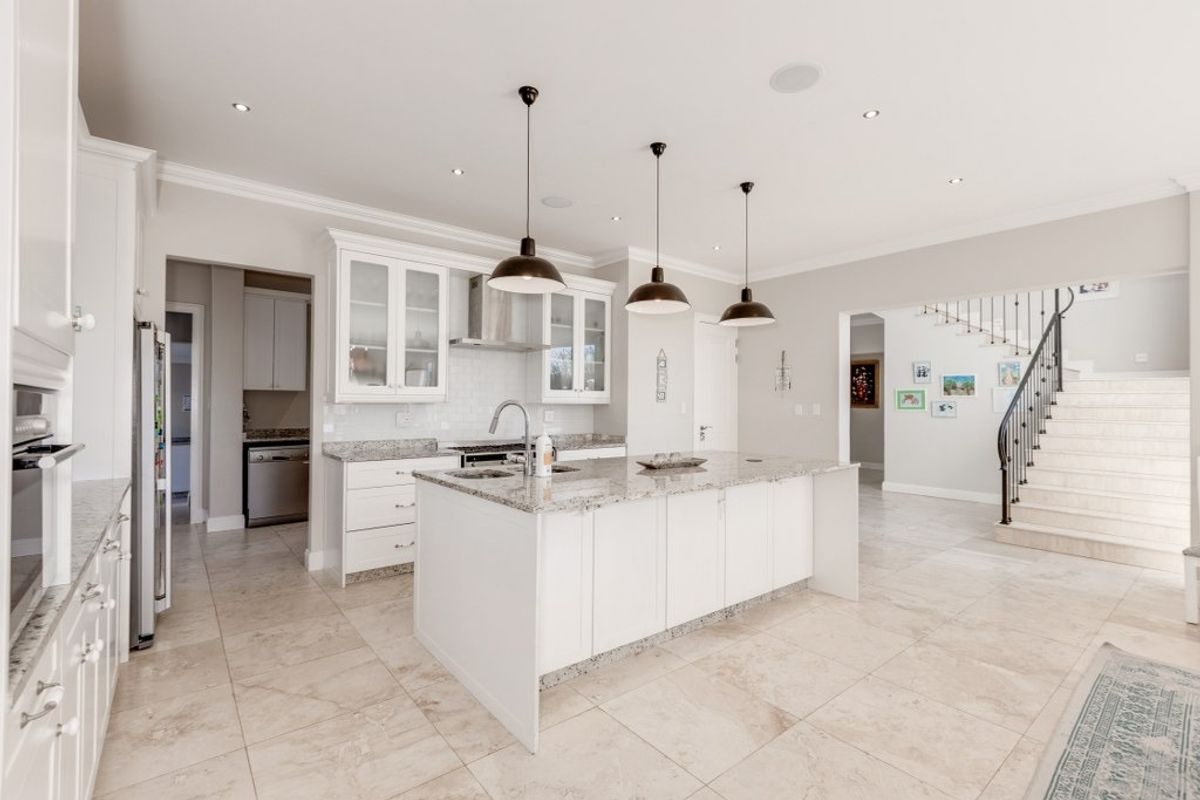 Modern kitchen featuring a central island with granite countertops, white cabinetry, stainless steel extractor fan, and tiled flooring, adjacent to an elegant staircase.