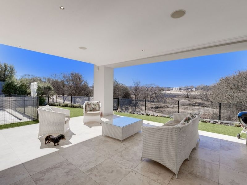 A spacious covered patio featuring high-quality travertine flooring, recessed lighting, and built-in ceiling speakers, overlooking a landscaped garden and open landscape.