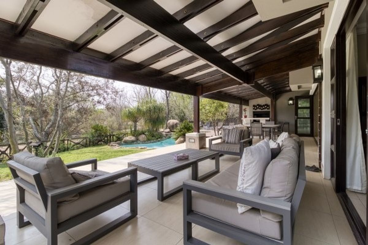Spacious covered patio featuring exposed timber beams, tiled flooring, and a built-in braai, overlooking a landscaped garden and pool area.