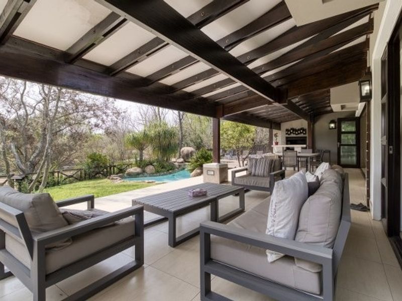 Spacious covered patio featuring exposed timber beams, tiled flooring, and a built-in braai, overlooking a landscaped garden and pool area.
