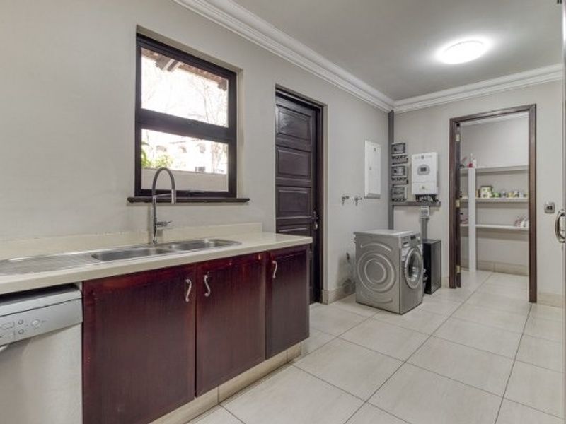 A functional laundry room featuring dark wood cabinetry, a stainless steel sink, tiled flooring, and direct access to a walk-in pantry or storage area.