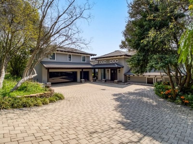 A well-maintained front exterior featuring a spacious paved driveway, double garage, and established landscaped gardens with mature trees.