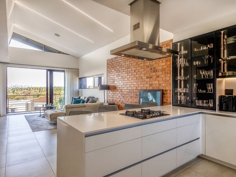 Modern open-plan living area featuring a sleek kitchen with island, gas hob, exposed brick wall, and seamless flow to a balcony with scenic views.