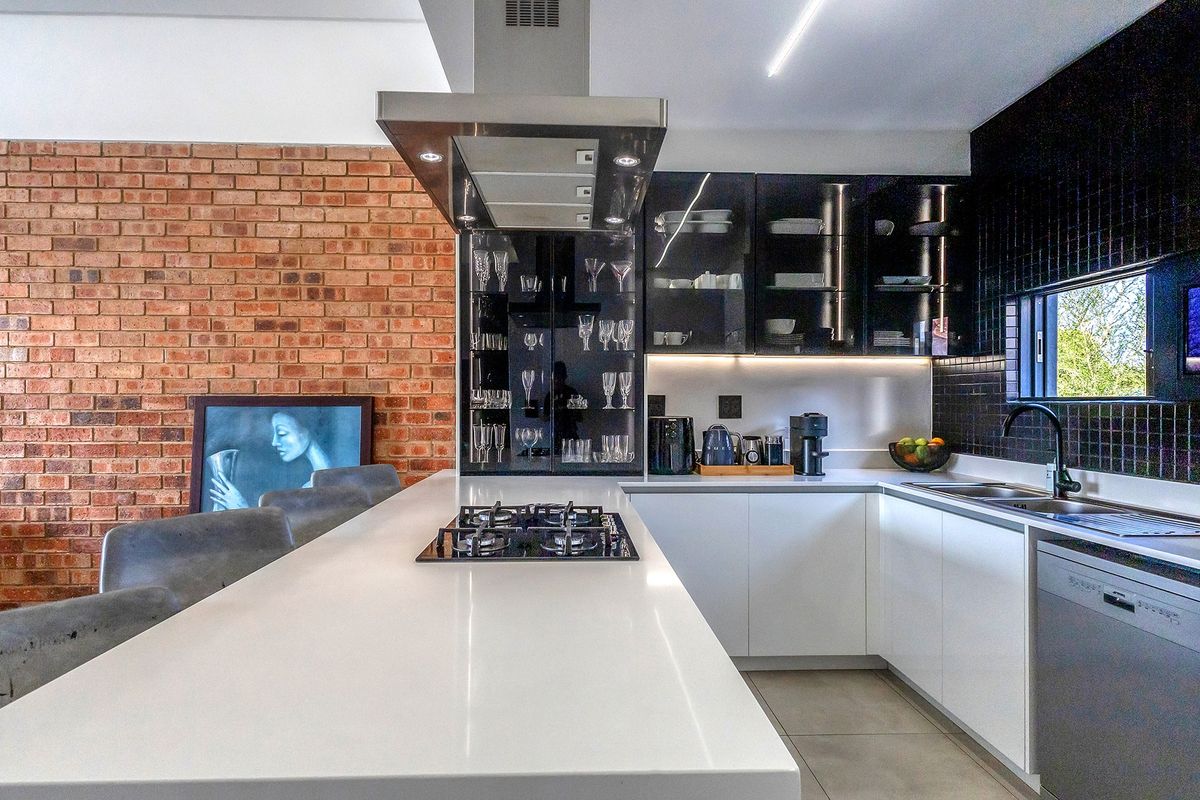 Modern kitchen featuring a central island with gas hob, sleek white cabinetry, quartz countertops, and a striking exposed brick feature wall.