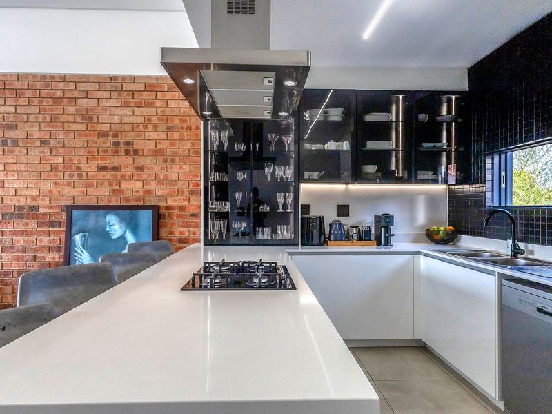 Modern kitchen featuring a central island with gas hob, sleek white cabinetry, quartz countertops, and a striking exposed brick feature wall.