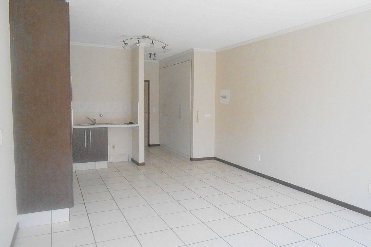 An empty open-plan living area featuring tiled flooring, a compact kitchenette with built-in cabinetry, and integrated storage cupboards.