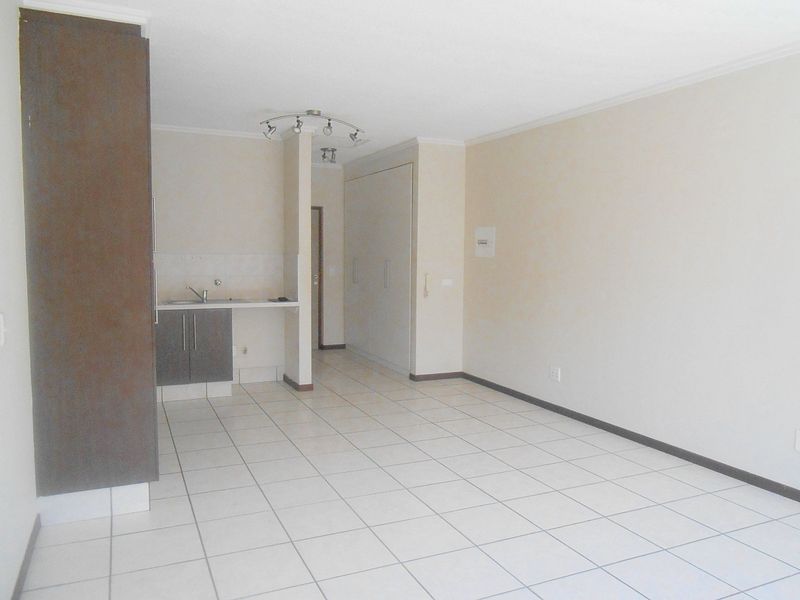 An empty open-plan living area featuring tiled flooring, a compact kitchenette with built-in cabinetry, and integrated storage cupboards.
