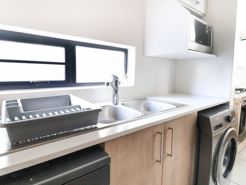 A modern kitchen featuring light wood-tone cabinetry, stone countertops, a double stainless steel sink, and integrated microwave, complemented by sleek black window frames.