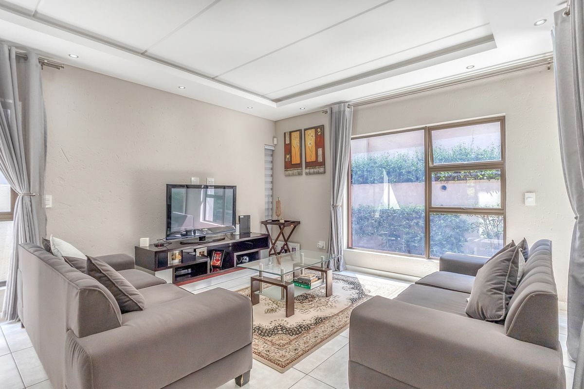 A well-lit living room featuring a recessed ceiling with downlights, tiled flooring, and large aluminium-framed windows overlooking the exterior.