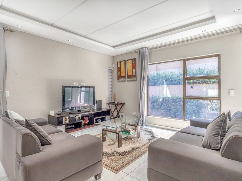 A well-lit living room featuring a recessed ceiling with downlights, tiled flooring, and large aluminium-framed windows overlooking the exterior.