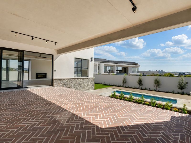 A spacious covered patio featuring herringbone brick paving, track lighting, and seamless access to the garden and swimming pool area.