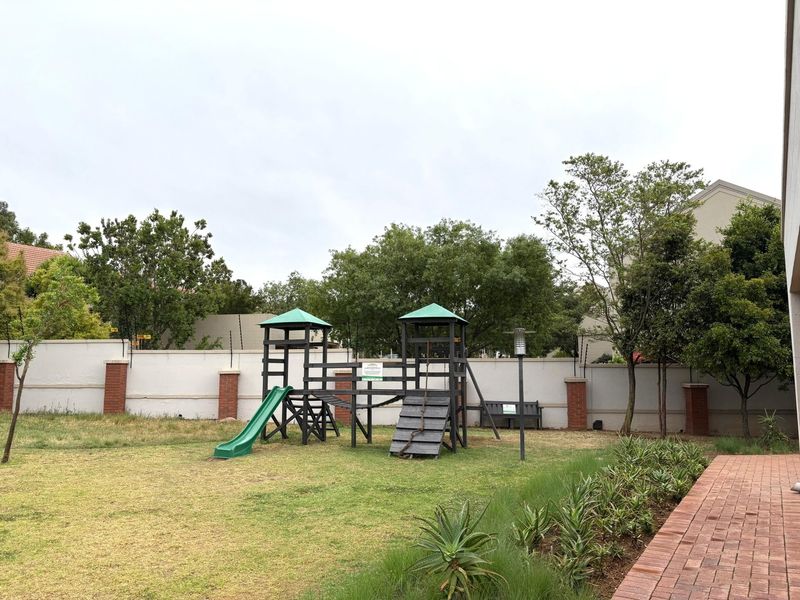 A well-maintained communal garden area featuring a children's play structure, paved pathways, and secure perimeter walling with electric fencing.