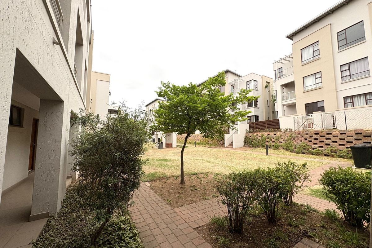 A view of the communal garden area between apartment blocks, featuring paved walkways, established landscaping, and a tiered retaining wall.