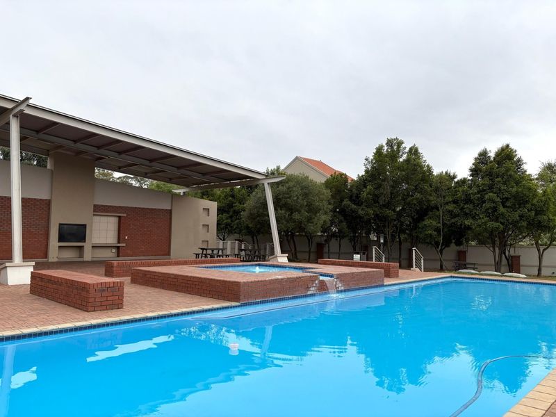 Communal pool area featuring a large swimming pool, raised brick spa with water feature, and a covered patio with built-in braai facilities.