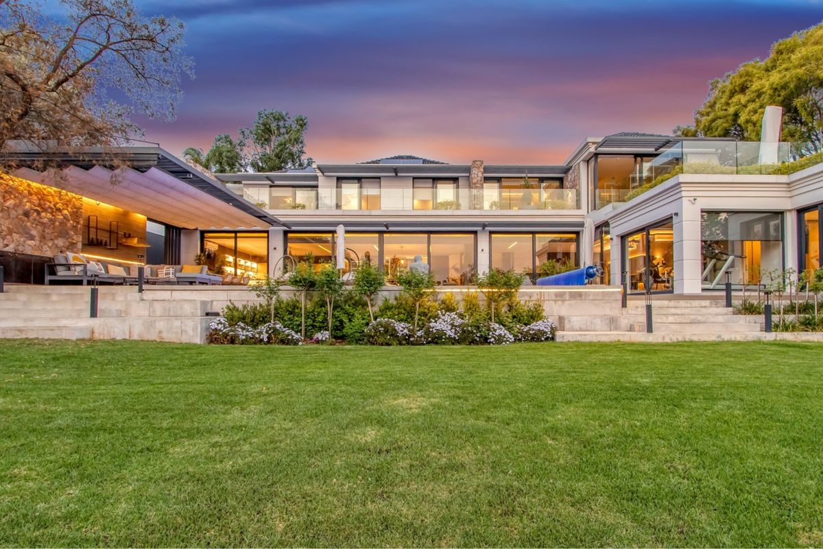 A stunning rear elevation of a modern luxury home featuring a spacious terrace, stone accents, glass balustrades, and expansive sliding doors overlooking a manicured garden.