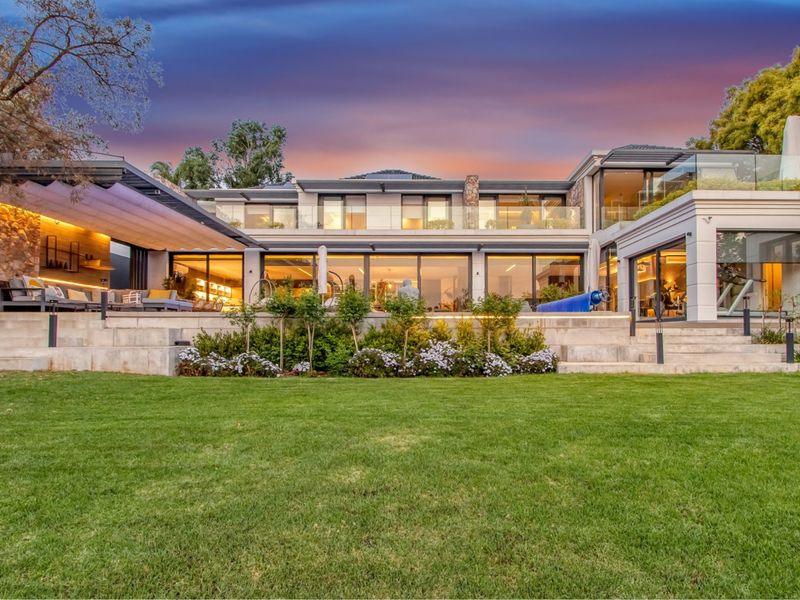 A stunning rear elevation of a modern luxury home featuring a spacious terrace, stone accents, glass balustrades, and expansive sliding doors overlooking a manicured garden.